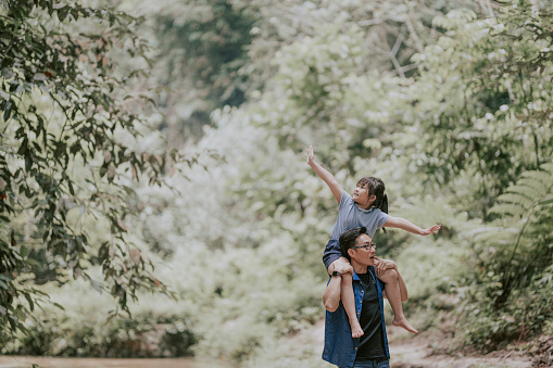 an asian chinese father carrying his daughter on shoulder in the jungle enjoying bonding time together at the river during weekend leisure time