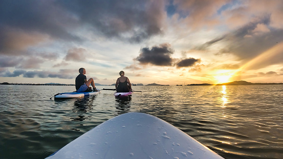 Two middle aged Women are sitting on paddle boards at sea watching a beautiful sunrise on the horizon.  Relaxing in nature image with copy space.  Filmed from a paddle boarder point of view on the Andaman sea, Thailand.