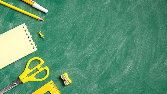 Back to school concept. Flat lay yellow school stationery on green chalkboard background. Top view ruler, scissors, paper note, pen, pencil, sharpener.