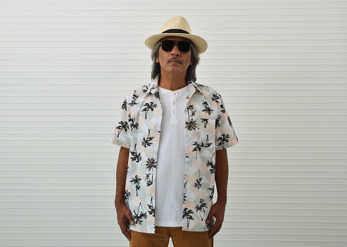 Confident senior traveler asian man wearing straw hat, sunglasses, summer shirt and brown shorts standing over white wall background, Business summer holiday concept