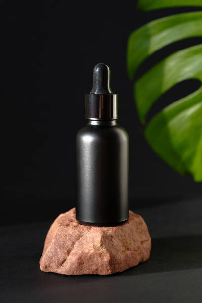 istock Black dropper bottle stands on stone and tropical palm leaf on black background. Premium beauty product packaging design mockup, herbal cosmetic. 1255334864