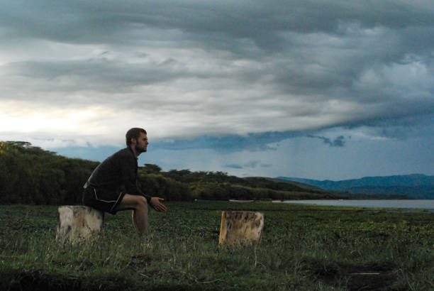 istock Side view man sitting on a log at an African lake staring forward on an overcast dramatic sky morning 1255338894