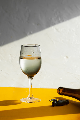 filled glass with water drops, dark glass wine bottle and corkscrew on a bright yellow background in natural light with sharp shadows, vertical image, summer bar menu