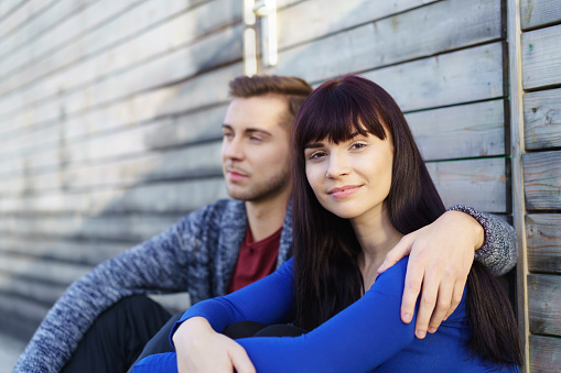 Attractive contented young woman sitting outdoors against a timber wall in a close embrace with her boyfriend turning to give the camera a warm friendly smile