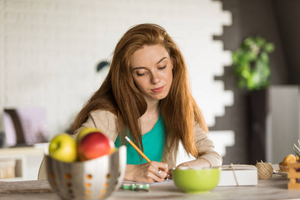Caring young woman sitting at the table in her home and writing a thoughtful letter to send with a gift box she put together during quarantine.