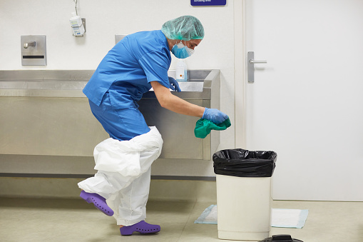 Side view of female nurse wearing scrubs, surgical mask, and bouffant cap removing protective suit and other disposable safeguards after use in trash bin.