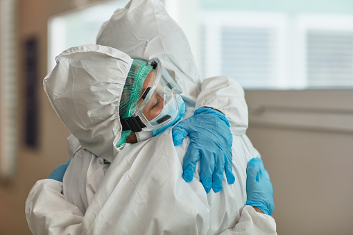 Male and female medical coworkers wearing protective suits, eyewear, and gloves hugging in support and recognition of hard work during COVID-19 pandemic.