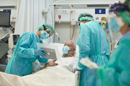 Senior male patient lying in hospital bed wearing multiple masks and communicating with family on digital tablet held by nurse in protective barrier sleeve.