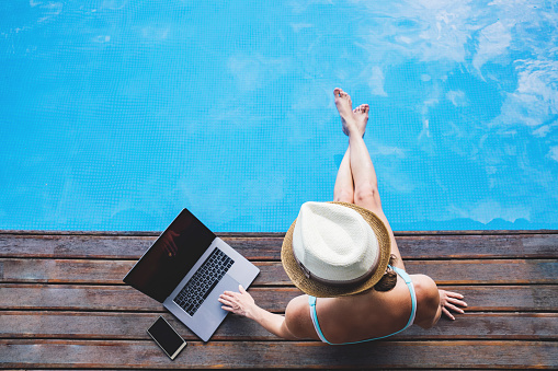 Young woman doing some digital on-line distant business, staying at home, sitting near water pool. Self isolation concept, working at home.