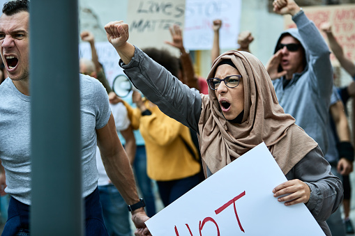 Large multi-ethnic group of people protesting against racism on the streets. Focus is on angry Middle Eastern woman shouting with raised fist.