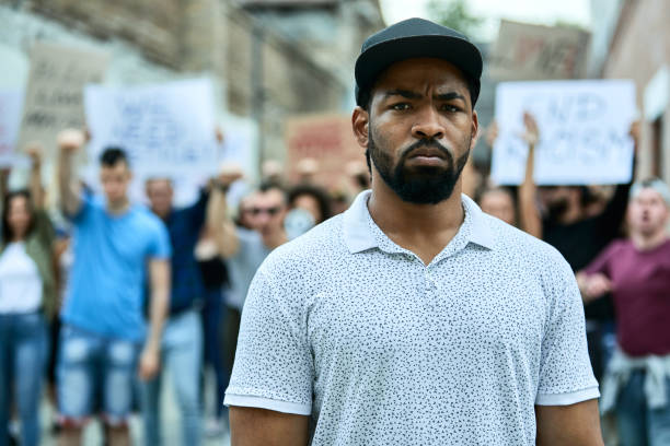 Young African American man on protests protest. Young black man participating in a protest for human rights and looking at the camera. Crowd of people is in the background. police brutality photos stock pictures, royalty-free photos & images