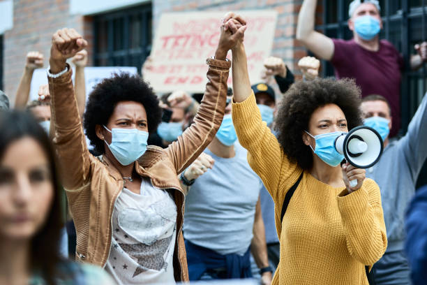 Black female activists holding hands while protesting with crowd of people during coronavirus pandemic. African American woman wearing protective face masks and holding hands while participating in demonstrations for human rights on the streets. unfairness photos stock pictures, royalty-free photos & images