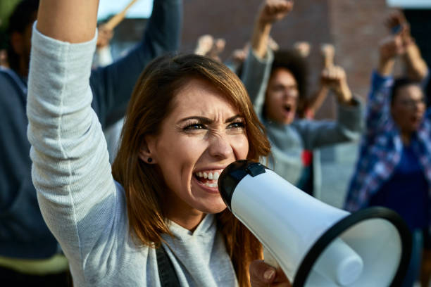istock Displeased woman shouting through megaphone while protesting with crowd of people on the streets. 1255363324