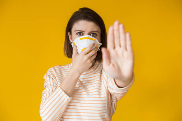istock Serious caucasian lady showing the stop sign while wearing a mask with filter on a yellow wall 1255371008