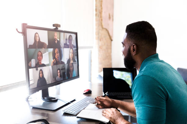 Man talking with colleagues during video call An African American businessman takes notes while discussing something with a team of colleagues during a video conference. virtual event photos stock pictures, royalty-free photos & images