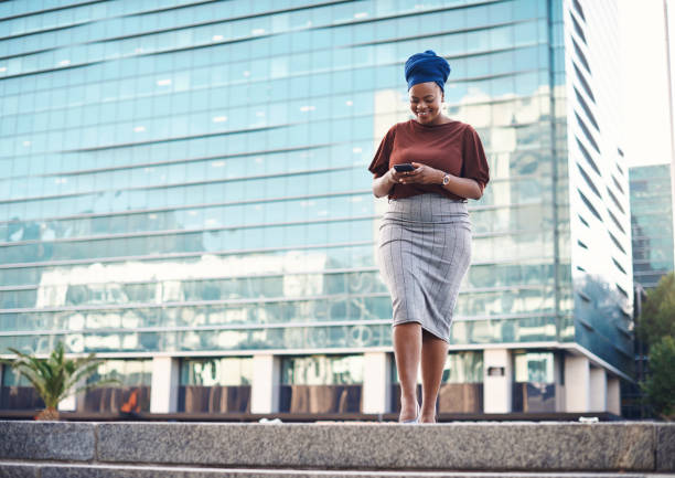 It's about time you stepped into the business spotlight Shot of a young businesswoman using a smartphone against a city background leanincollection stock pictures, royalty-free photos & images