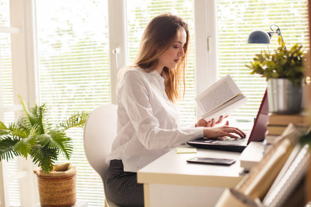 istock Young woman studying at her desk and typing in notes from a book she is holding 1255382566