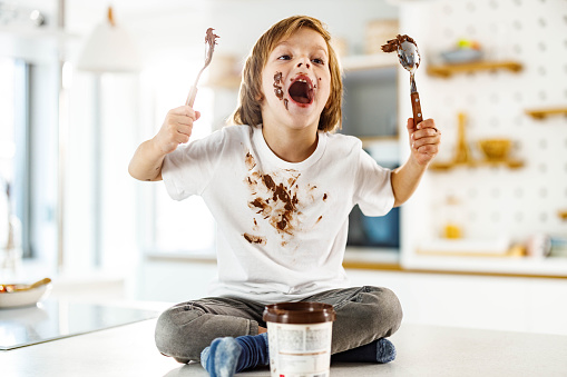 Messy little boy with stains having fun while eating chocolate spread in the kitchen.
