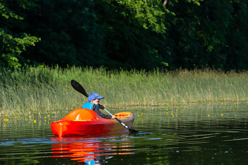 An almost four year old boy kayaking on a lake. He is paddling with his black paddle, and he is in an orange and yellow kayak. The boy is wearing a blue baseball cap and life jacket.