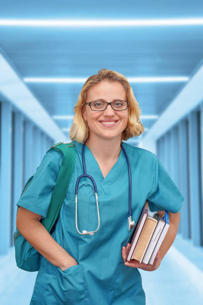 trainee surgeon with eyeglasses in a blue uniform with books and a bag on the background of a blurred corridor of the clinic. medical doctor over blue health care background. education concept. - medical student healthcare and medicine book education imagens e fotografias de stock