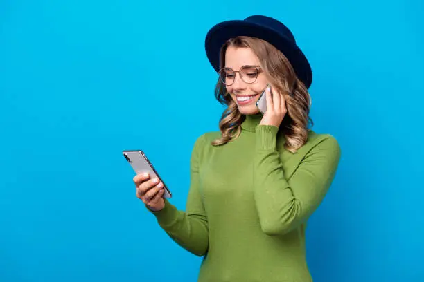 Photo of Photo of cheerful funny curly lady hold telephone popular blogger writing new post talking second phone listen advert offer wear specs hat green turtleneck isolated blue color background