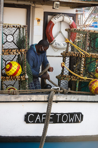 Cape Town, South Africa – July 4: Unidentified fisherman secures boats with rope while wearing a mask in Kalk Bay Harbour