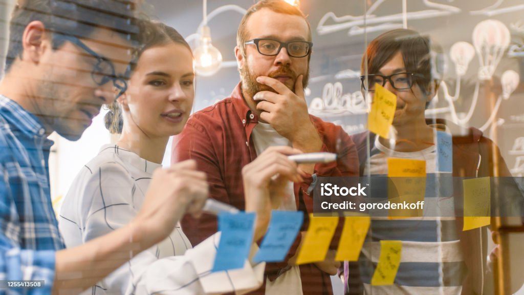 Diverse Team of Young Developers Brainstorming, They Have Discussion and Use Glass Board and Sticky Papers to Conceptualize Their New Plan. Creative People in Stylish Office Environment. Contemplation Stock Photo