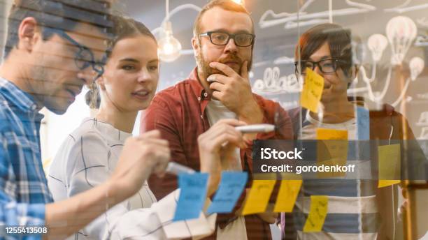 Diverse Team Of Young Developers Brainstorming They Have Discussion And Use Glass Board And Sticky Papers To Conceptualize Their New Plan Creative People In Stylish Office Environment Stock Photo - Download Image Now
