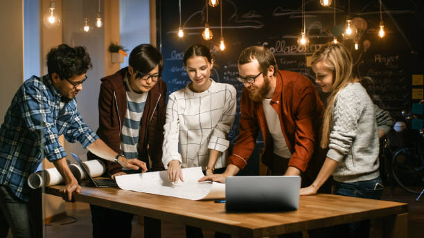 istock Late at Night Team of Young Creative Developers at their Meeting Table Spread Blueprint and Get Down to Work. Stylish People in Trendy Office Environment. 1255413699