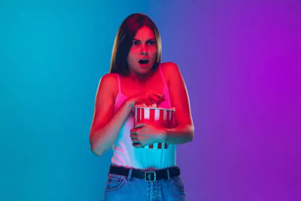 Shocked, scared, eating popcorn. Caucasian young woman's portrait on gradient background in neon light. Beautiful female model. Concept of human emotions, facial expression, sales, ad, movie, cinema.