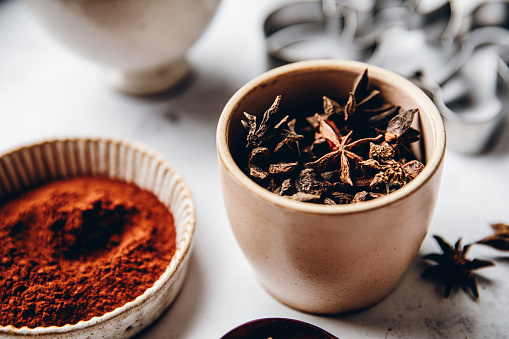 Traditional Christmas spices in a bowl with cocoa powder and cutter in background for preparing Christmas cookies. Star anise spice in a ceramic bowl.