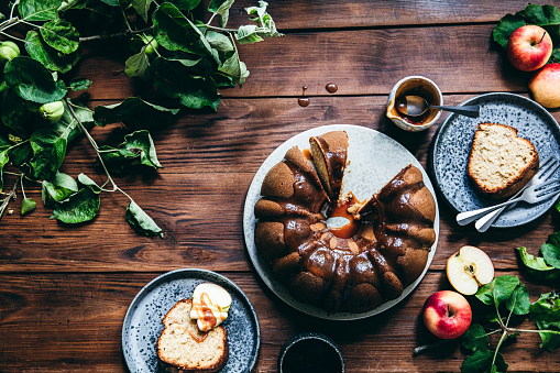 Top view of a freshly made apple bundt cake with caramel sauce on wooden table. Slice of apple cake served in a plate with freshly harvested apples on a wooden table.