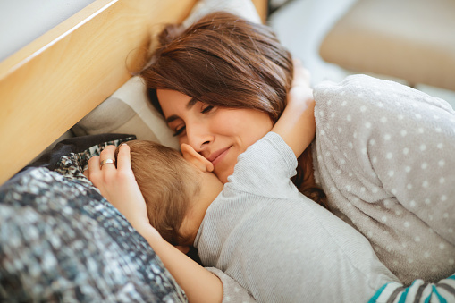 Smiling mother holding her sleeping toddler in arms, lying in bed in pajamas