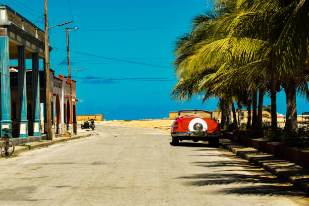 An old American car with its roof down parked on a coastal city street lined with palm trees in Gibara, Cuba