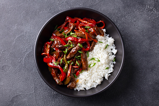 Beef and vegetables stir fry with white rice in a black bowl. Grey background. Top view.