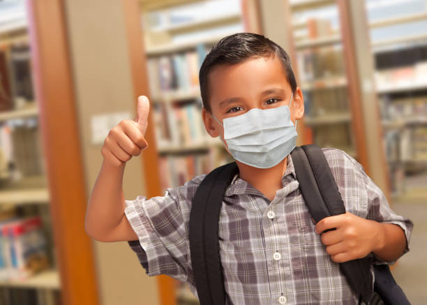 図書館で親指とバックパックを持つフェイスマスクを着用したヒスパニック学生の男の子 - thumbs up book smiling student ストックフォトと画像