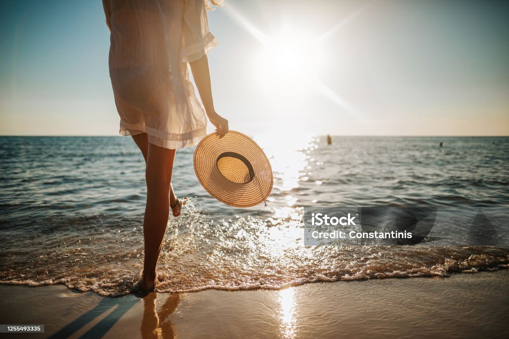 Woman's legs splashing water on the beach Close-up of young woman in white sun dress and with hat in hand walking alone on sandy beach at summer sunset, splashing water in sea shallow Beach Stock Photo