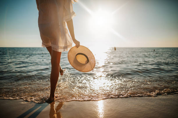Woman's legs splashing water on the beach Close-up of young woman in white sun dress and with hat in hand walking alone on sandy beach at summer sunset, splashing water in sea shallow island vacation stock pictures, royalty-free photos & images