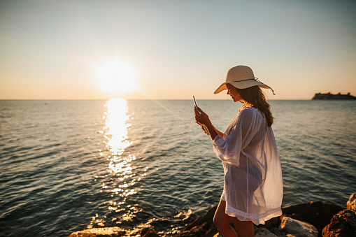 Beautiful young woman in white sun dress and with hat taking selfie with her digital tablet on the beach at beautiful summer sunset above the sea
