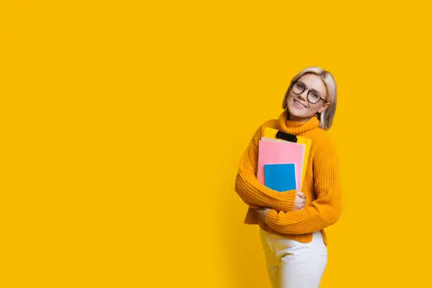 Blonde student with eyeglasses smiling at camera and holding some books while posing on a yellow wall with freespace
