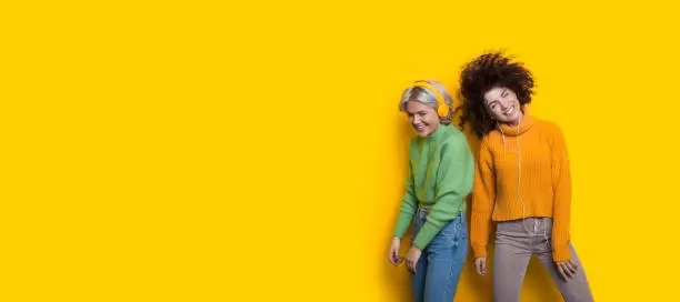 Photo of Curly haired friends listening to music using headphones on a yellow freespace while dancing happily