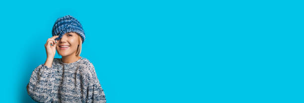 istock Nice woman with blue hat and blonde hair touching it while smiling at camera on a blue wall with empty space 1255509097