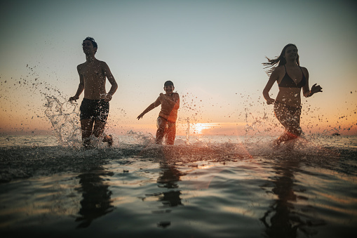 Brothers and sister having fun at sea, cheerfully running in sea shallow at sunset, smiling and splashing