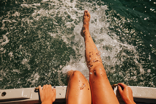 Close-up of young woman's legs sitting on a boat deck and splashing on the water
