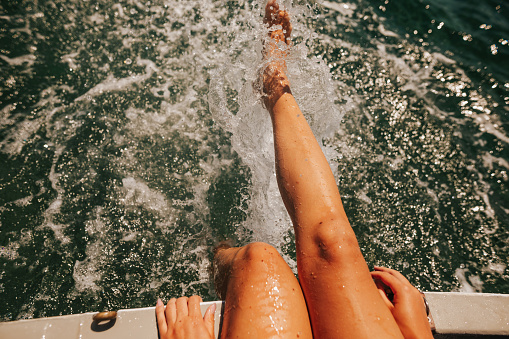 Close-up of young woman's legs sitting on a boat deck and splashing on the water