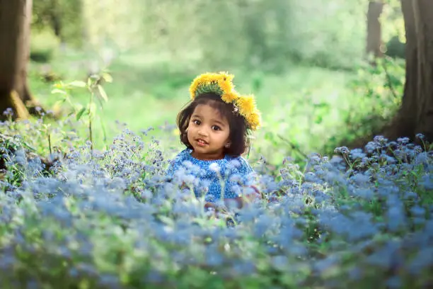 A small girl with dandelion wreath is sitting on field of forget me not blue flowers