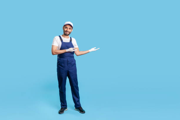 istock Full length cheerful workman in blue overalls, cap and protective gloves standing, showing empty space 1255536568