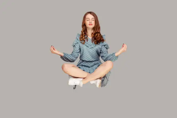 Hovering in air. Calm peaceful relaxed girl ruffle dress levitating with mudra gesture hands up, closed eyes, meditating sitting in yoga position. indoor studio shot isolated on gray background