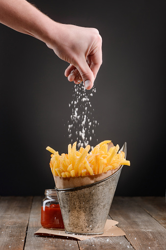 Hand adding a pinch of salt into French fries. Yummy snack served with a jar of ketchup sauce.