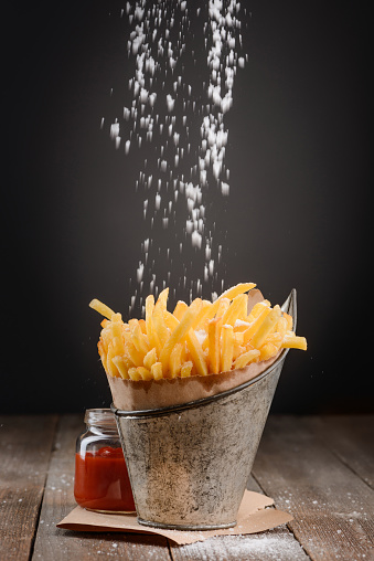 Pinch of salt falling on French fries. Whole bucket of favorite crispy snack served with ketchup on rustic wooden table.